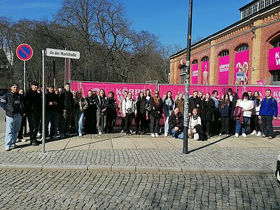 Eine große Gruppe von Jugendlichen und jungen Erwachsenen steht im Halbkreis vor einem teils rosa angemalten Gebäude.