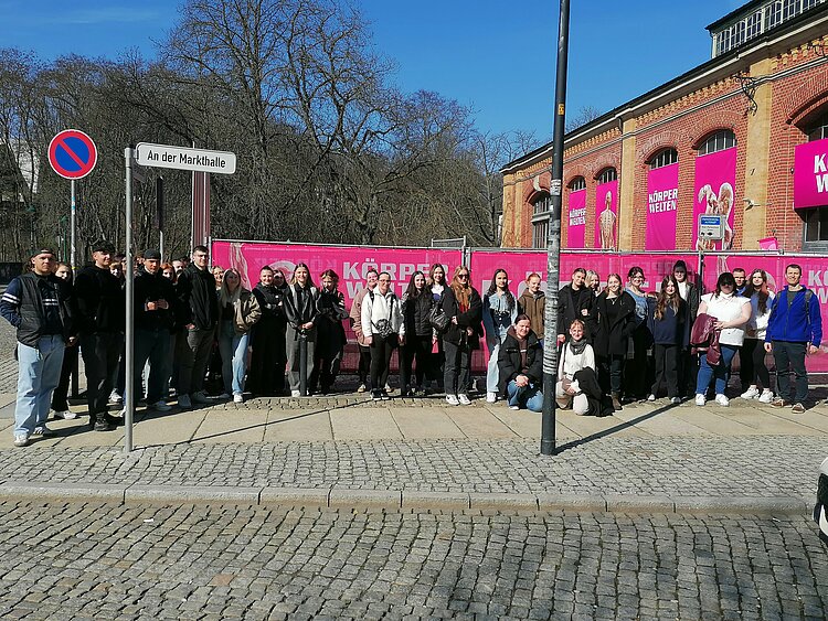 Eine große Gruppe von Jugendlichen und jungen Erwachsenen steht im Halbkreis vor einem teils rosa angemalten Gebäude.