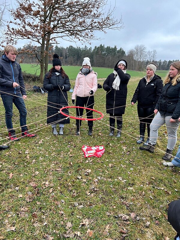 Mehrere Schüler*innen stehen in einem Kreis und halten jeweils das Ende eines dicken Fadens fest. In der Mitte laufen alle Fäden zusammen, denn dort sind sie an einem größeren roten Plastikring befestigt. Die Szene spielt sich auf einer Wiese ab, im Hintergrund ist ein Wald zu sehen.