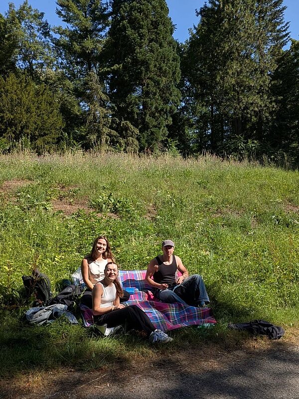 Drei Personen sitzen auf einer Decke in einem grünen, leicht hügeligen Bereich. Sie lächeln in die Kamera, während sie sich entspannen. Im Hintergrund sind Bäume und ein Wald zu sehen.