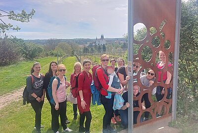Eine Gruppe von Frauen, die Rucksäcke tragen, steht vor eine rostigen Stelle, aus der Kreise ausgeschnitten sind. Einige Frauen schauen durch diese Löcher in die Kamera. Eine Frau hat ein Kind einem Tragetuch. Hinter der Gruppe befindet sich ein Weinberg, dahinter Umrisse einer Stadt.