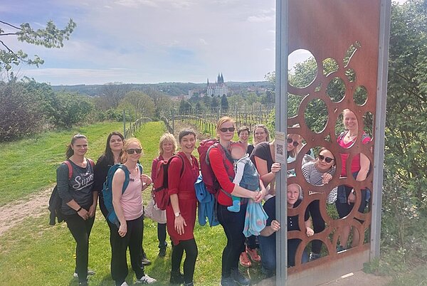 Teamevent der Euro Akademie Eine Gruppe von Frauen, die Rucksäcke tragen, steht vor eine rostigen Stelle, aus der Kreise ausgeschnitten sind. Einige Frauen schauen durch diese Löcher in die Kamera. Eine Frau hat ein Kind einem Tragetuch. Hinter der Gruppe befindet sich ein Weinberg, dahinter Umrisse einer Stadt.