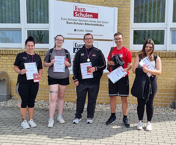Wikinger-Sportfest: Pflegeazubis erobern die nordische Arena Fünf junge Erwachsene stehen vor einem Schulgebäude und halten Urkunden sowie Medaillen. Sie posieren stolz für ein Gruppenfoto.