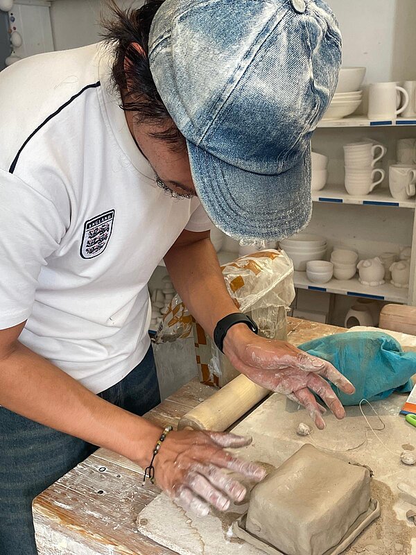 Junger Mann mit Jeans-Cap formt an einem Arbeitstisch mit beiden Händen eine rechteckige Schale aus Ton. Im Hintergrund stehen Regale mit Keramikgefäßen.