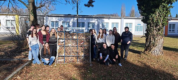 Auf einer laubbedeckten Wiese vor einem flachen weißen Gebäude steht ein selbstgebautes Insektenhotel, gefüllt mit Laub; drumherum steht eine Gruppe Schüler und Schülerinnen.