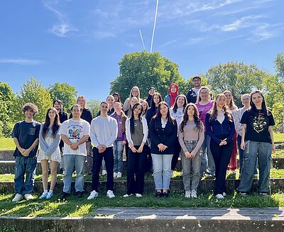 Absolventinnen und Absolventen der Euro Akademie Aschaffenburg stehen draußen, bei herrlichem Sommerwetter und blauem Himmel, zusammen und posieren für ein Gruppenfoto.