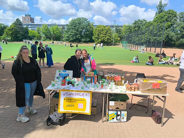 Schnitzeljagd, Tombola und Volleyball-Turnier Zwei weibliche Auszubildende posieren gut gelaunt und lächelnd für ein Foto, sie stehen am Tombolatisch, der mit allerlei Preisen bestückt ist.