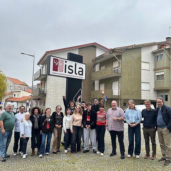 Eine Gruppe von Teilnehmenden der Businet-Konferenz posiert vor einem Gebäude mit der Aufschrift „Isla Instituto Politécnico de Gestão e Tecnologia“. Die Gruppe ist gemischt, Männer und Frauen unterschiedlichen Alters; einige heben die Hände, andere lächeln. Das Gebäude im Hintergrund wirkt modern, der Himmel ist bedeckt.