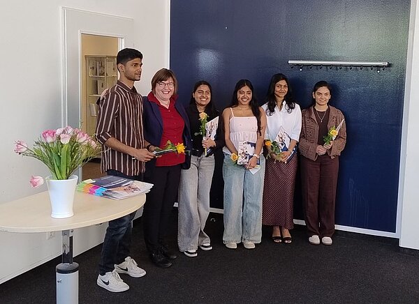 Sechs Personen stehen in einem Raum mit blauer Wand und einem runden Tisch, auf dem ein Blumenstrauß in einer weißen Vase steht. Fünf der Personen halten Zertifikate und Blumensträuße. Die Gruppe wirkt stolz und glücklich, möglicherweise bei einer offiziellen Feier oder Zeremonie. Eine Person trägt ein gestreiftes Hemd, eine andere einen gemusterten Blazer.