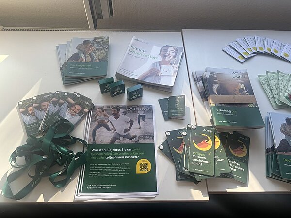 Ein Tisch mit verschiedenen Informationsmaterialien und Give-aways. Dazu gehören Broschüren, Flyer, Visitenkarten, ein QR-Code, kleine grün-weiße Schachteln und Lanyards mit Aufdrucken wie 'AOK PLUS'. Die Materialien sind ordentlich arrangiert und wirken einladend.