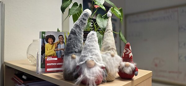Vier weihnachtlich dekorierte Wichtelfiguren mit langen, spitzen Mützen in Grau-, Weiß- und Rottönen stehen auf einer hellen Holzoberfläche. Im Hintergrund ist eine grüne Pflanze mit großen Blättern zu sehen. Neben den Wichteln steht ein aufgestelltes Informationsheft der Euro-Schulen mit dem Titel 'Deutsch lernen'. Die Szene vermittelt eine festliche Atmosphäre in den Räumlichkeiten der Euro-Schulen und Euro Akademien in Dresden, Meißen und Riesa.