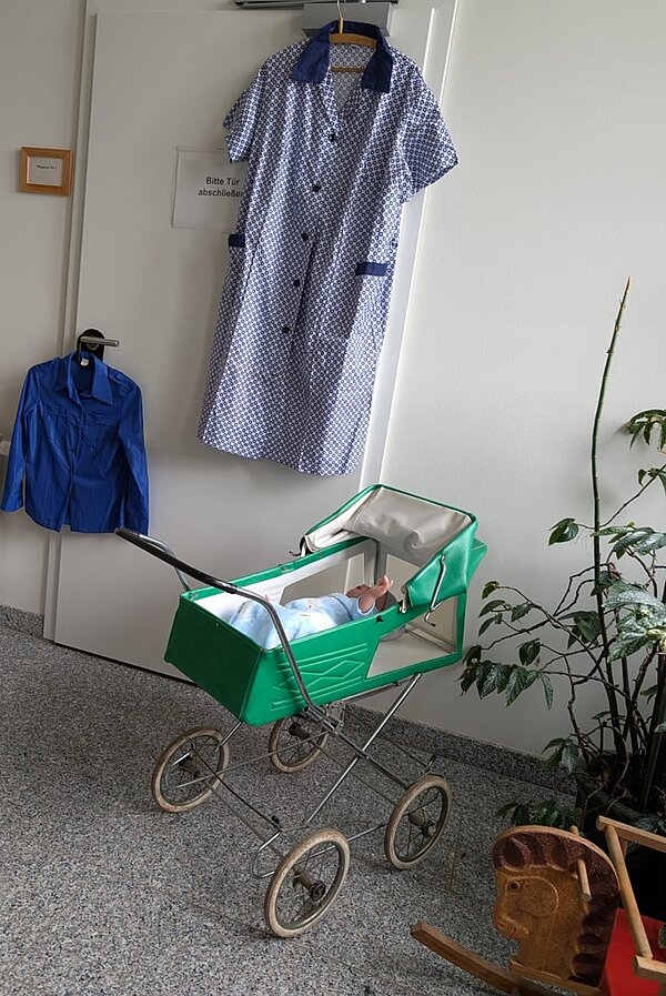 Ein grüner Kinderwagen mit cremefarbenem Innenfutter steht auf einem grauen Boden. Im Kinderwagen liegt eine Puppe mit heller Kleidung. Im Hintergrund hängt ein blau-weiß gemustertes Kleid mit kurzen Ärmeln an einem Kleiderbügel an einer weißen Tür. Daneben hängt ein blaues Hemd. Rechts im Bild steht eine grüne Zimmerpflanze mit langen, schmalen Blättern. Unten rechts ist ein Teil eines Holzspielzeugs zu sehen.