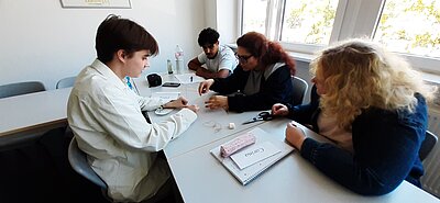 Vier Personen sitzen an einem Tisch im Klassenzimmer und arbeiten gemeinsam an einer Übung mit Fäden, Stäbchen und Marshmallows.