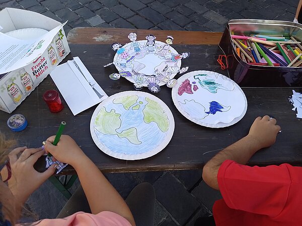 Kinder malen auf Papptellern Weltkarten aus, daneben ausgeschnittene Papierfiguren und Buntstifte in einer Metallbox.