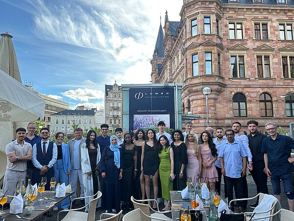 Abschlussfeier mit Zeugnisübergabe Eine große Gruppe festlich gekleideter junger Menschen und einige Erwachsene posieren gemeinsam im Außenbereich eines Restaurants vor einem historischen Gebäude. Die Stimmung ist feierlich, die Tische sind mit Getränken und Gedecken vorbereitet.