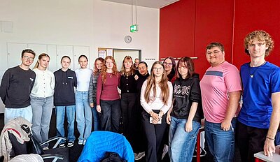 Schülerinnen und Schüler aus der Slowakei posieren an der Euro Akademie Bamberg für ein Gruppenbild. Alle sind leger gekleidet in sportlichen Pullovern, ein Schüler in einem rosa T-Shirt, ein anderer in blauem T-Shirt, im Hintergrund sieht man eine dunkelrot gestrichene Wand. Alle lächeln gut gelaunt in die Kamera. 