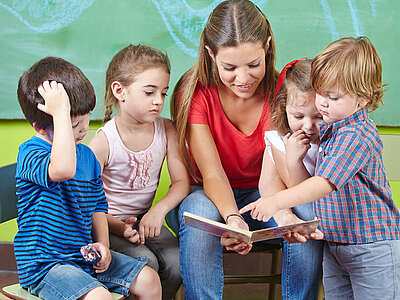Eine Frau mit rotem Top hält ein Kinderbuch in den Händen. Links und rechts von ihr sitzen Kinder, die mit ihr gemeinsam in das Buch schauen.