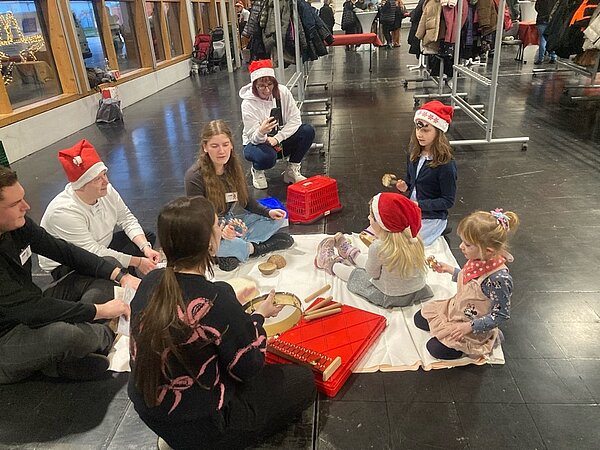 Eine Gruppe von Erwachsenen und Kindern sitzt im Kreis auf dem Boden und spielt mit verschiedenen Musikinstrumenten. Alle tragen Weihnachtsmützen, und im Hintergrund sind Kleiderständer zu sehen.
