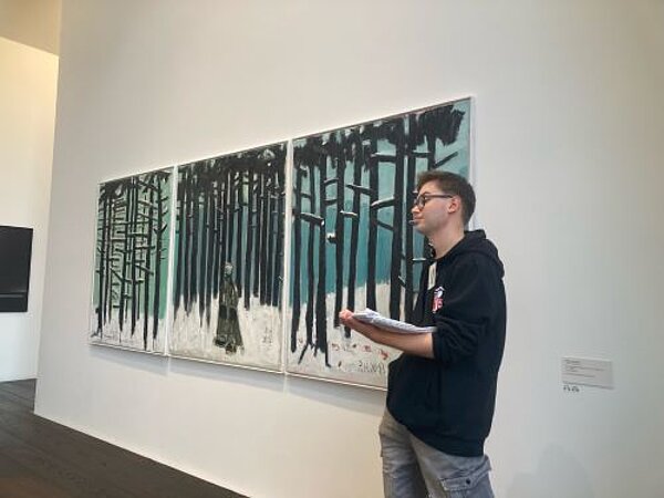 Ein junger Mann in einem schwarzen Hoodie und Brille steht vor einem großen, abstrakten Kunstwerk, das Bäume und eine winterliche Landschaft darstellt. Er hält ein Blatt Papier in der Hand und scheint das Kunstwerk zu erklären.