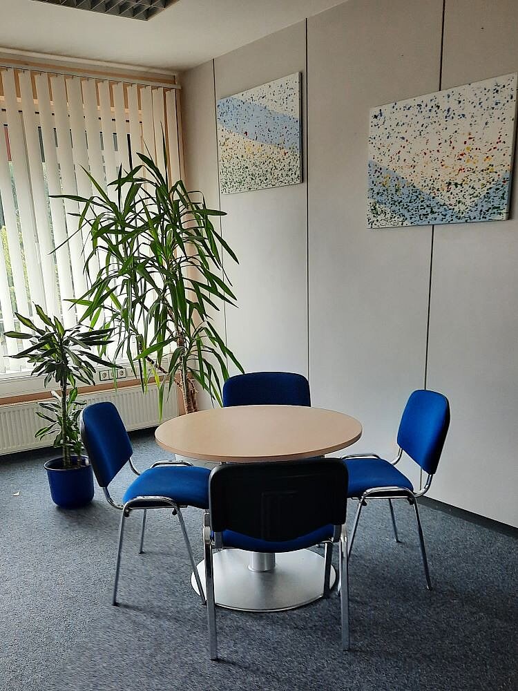 Ein kleiner Besprechungsraum mit einem runden Tisch und vier blauen Stühlen. Im Hintergrund stehen zwei bunte Gemälde an der Wand. Hinter der Tischgruppe befindet sich ein großer Grünpflanze in einem Topf, die vor einem Fenster mit Lamellen steht.