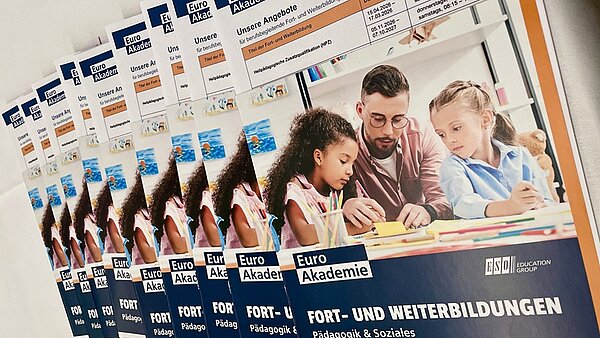 Mehrere Broschüren liegen leicht versetzt übereinander. Auf dem Titelbild ist ein Erwachsener mit zwei Kindern an einem Tisch zu sehen, alle blicken nach vorn. Die Broschüren tragen den Titel 'FORT- UND WEITERBILDUNGEN' und zeigen das Logo 'Euro Akademie'. Die Farbgestaltung ist überwiegend blau und weiß, mit orangefarbenen Akzenten. Im oberen Bereich sind weitere Textfelder und Logos sichtbar.