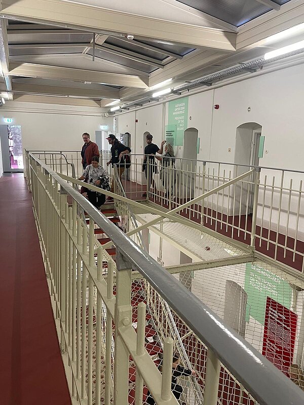 Abschlussklasse der Erzieher*innen besucht Kaßberg-Gefängnis in Chemnitz Blick auf die obere Etage des Kaßberg-Gefängnisses mit Besuchern auf dem Gittergang. An den Wänden hängen Informationstafeln, die Zellentüren sind sichtbar.