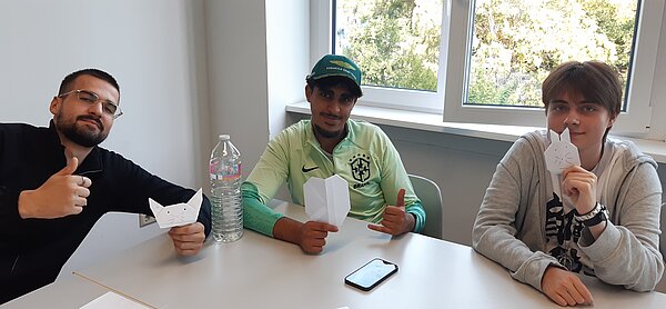 „Die Neuen“ sind da – Euro Akademie Mainz begrüßt neue Klasse der Fremdsprachenkorrespondenten Drei Personen sitzen nebeneinander, zeigen ihre Origami-Figuren und lächeln in die Kamera, zwei heben zusätzlich den Daumen hoch.