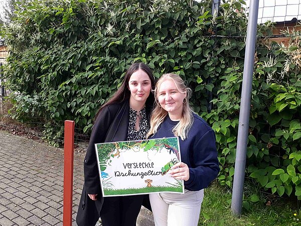 Zwei Auszubildende posieren mit ihrem Schild versteckte Dschungeltiere lächelnd für ein Foto.