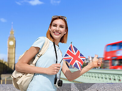 Eine lächelnde Frau steht vor einer berühmten Londoner Sehenswürdigkeit, dem Big Ben, und hält eine britische Flagge in der Hand. Sie trägt eine Sonnenbrille auf dem Kopf, ein hellblaues T-Shirt und einen beigen Rucksack. Im Hintergrund ist ein roter Doppeldeckerbus zu sehen, der über eine Brücke fährt.