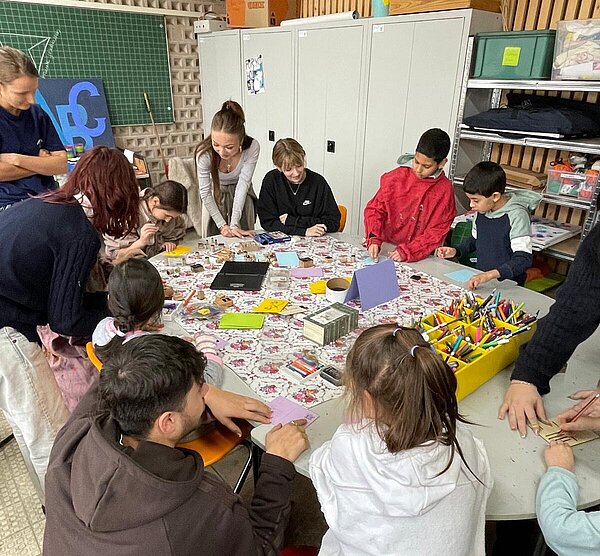 Gruppe von Kindern und Anleitenden arbeitet an einem großen Bastel- und Drucktisch mit Stempeln, Papieren und Stiften im Klassenraum.