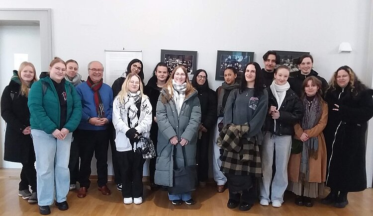 Eine Gruppe von Auszubildenden der Euro Akademie Bamberg steht in einem hellen Raum des Amtsgerichts Fürth vor einer weißen Wand. Im Hintergrund hängen zwei gerahmte Fotografien, die Szenen aus den Nürnberger Prozessen zeigen. Die Personen tragen winterliche Kleidung wie Mäntel, Schals und Jacken in gedeckten Farben. Der Boden besteht aus hellem Holz. Die Gruppe steht dicht beieinander und blickt in Richtung Kamera. Das Bild dokumentiert den Besuch der Auszubildenden bei der Ausstellung zu den Nürnberger Prozessen.