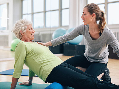 Zwei Personen sind in einer Physiotherapie-Stunde in einem hellen Raum mit großen Fenstern und blauen Übungsmatten. Eine Person hilft der anderen bei einer Übung.