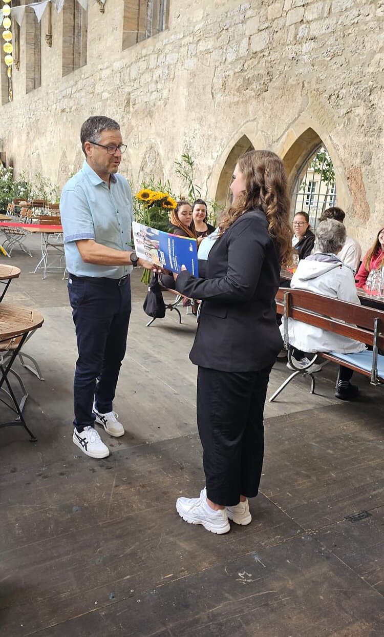 Feierliche Zeugnisübergabe in der Barfüßerruine Ein Mann in hellem Hemd und dunkler Hose überreicht einer jungen Frau in schwarzer Kleidung ein Zertifikat und einen Strauß mit Sonnenblumen. Beide stehen in einem Innenhof mit rustikalem Steinmauerwerk und Holzbänken im Hintergrund, auf denen Personen sitzen.