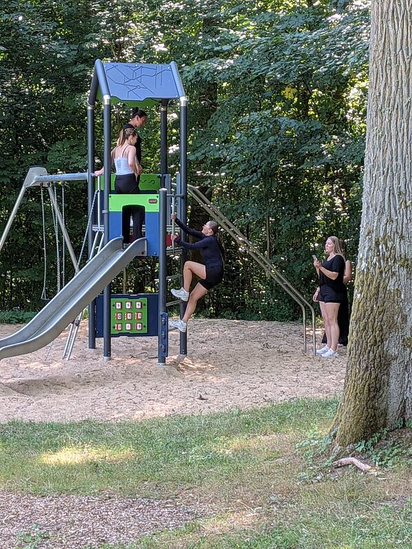 Zwei Personen stehen auf einem Spielplatzgerät mit Rutsche und Kletterelementen. Eine andere Person klettert die Leiter hinauf, während zwei weitere daneben stehen und die Szene mit dem Handy fotografieren. Im Hintergrund sind Bäume zu sehen.