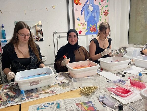 Drei Personen sitzen an einem Tisch und arbeiten an kreativen Projekten. Vor ihnen stehen weiße Kunststoffwannen mit farbigen Flüssigkeiten in Rot, Blau und Gelb. Der Tisch ist mit Zeitungen ausgelegt, und im Hintergrund hängen bunte Bilder an der Wand.