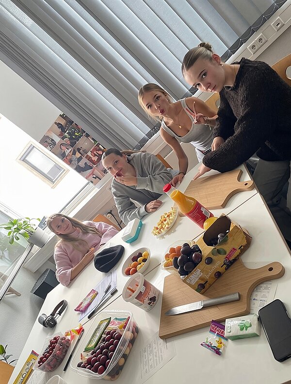 Vier junge Auszubildende sitzen an einem rechteckigen Tisch in einem hellen Raum und posieren für ein Foto. Auf dem Tisch liegen verschiedene Lebensmittel, darunter Obst wie Trauben, Pflaumen und Aprikosen, eine Packung Saft, ein Messer, ein Schneidebrett mit Apfelstücken, eine Schachtel Kaugummi und eine Packung Süßigkeiten. Im Hintergrund stehen ein grüner Blumentopf und mehrere Steckdosenleisten auf der Fensterbank. Sie tragen Freizeitkleidung und blicken in Richtung Kamera. An der Wand hängen mehrere Fotos oder Poster.