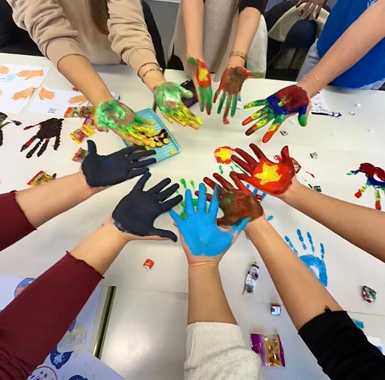 Eingängiges Projekt zum Thema Hygiene Eine Gruppe von Menschen halten ihre bunt angemalten Hände mit den Handflächen nach oben in einem Kreis aneinander. Darunter liegt ein weißes Plakat, auf dem bunte Handabdrücke zu sehen sind.