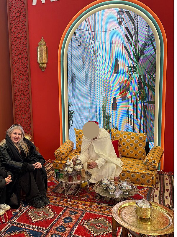 Schülerin sitzt lächelnd bei einer marokkanischen Teezeremonie; ein Mann in traditioneller Kleidung sitzt auf einem gelben Sofa und schenkt Tee aus; auf dem Boden und an der Wand sind rote Teppiche