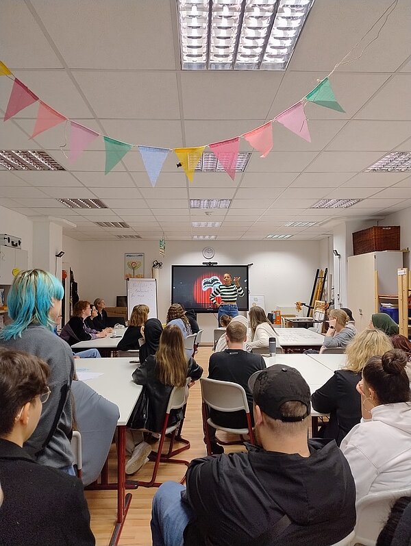 Studierende der Fachschule gestalten eindrucksvolle Ausstellung Die weibliche Lehrkraft steht im Klassenraum vor den Studierenden und gestikuliert erklärend. Die Studierenden hören aufmerksam zu. An der Zimmerdecke hängt eine bunte Wimpelkette.