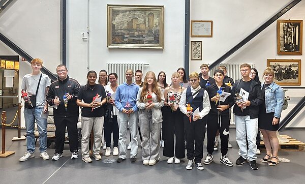 Herzlicher Abschied, freudiger Anfang: Pflege-Absolvent*innen geehrt und neue Auszubildende willkommen geheißen Gruppenfoto von jungen Erwachsenen, die lächelnd vor einer Wand stehen, an der ein Gemälde und historische Fotos hängen. Die Personen halten Zertifikate und kleine Geschenke in den Händen.