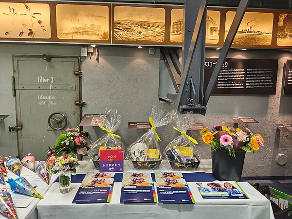 Herzlicher Abschied, freudiger Anfang: Pflege-Absolvent*innen geehrt und neue Auszubildende willkommen geheißen Ein Tisch ist mit Blumensträußen, Geschenkpaketen, Informationsbroschüren und kleinen Überraschungen dekoriert. Im Hintergrund sind historische Fotos und eine Metalltür mit der Aufschrift „Filter 1“ und „Lebensgefahr nicht öffnen“ zu sehen. Der Raum wirkt wie ein Industriegebäude.