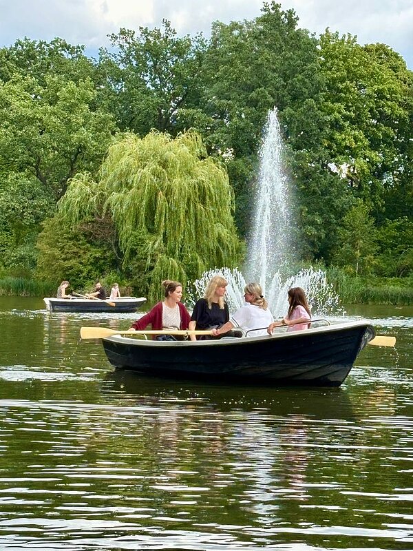 Zwei Gruppen von Menschen sitzen in Ruderbooten auf einem See und genießen die Zeit auf dem Wasser. Im Hintergrund ist ein großer Springbrunnen zu sehen, der Wasser in die Luft spritzt. Um den See herum sind Bäume und grüne Landschaft zu sehen.
