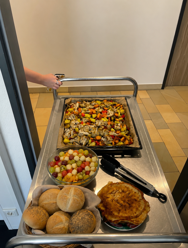 Ein Servierwagen steht in einem Flur. Auf dem oberen Blech liegt ein Blech mit gebackenem Gemüse und Hähnchenteilen, darunter Paprika, Zwiebeln und Kartoffeln in verschiedenen Farben. Daneben stehen eine Schale mit Brötchen, eine Schale mit Obstsalat aus Melonen und Trauben sowie ein Stapel Kartoffelpuffer. Eine Hand hält den Griff des Wagens, eine Zange liegt auf dem Tablett.