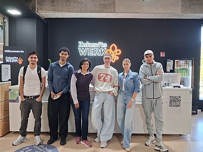 Vor einer weißen Theke stehen sechs Personen für ein Gruppenbild. An der schwarzen Wand hinter ihnen steht in Leuchtschrift "Zukunftswerk" und daneben ist ein orangefarbenes Logo abgebildet. Rechts hinten steht ein Kühlschrank mit Getränken.
