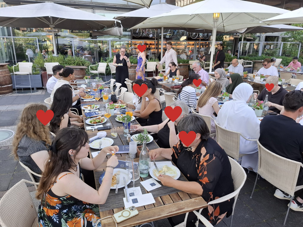 Große Gruppe sitzt gemeinsam an gedeckten Tischen auf einer Restaurantterrasse und isst zusammen.