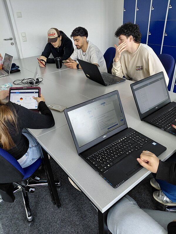 Drei Personen mit Laptops und Handys sitzen konzentriert an einem Tisch; gegenüber sitzt eine Person mit Tablet; im Vordergrund sind zwei weitere Laptops zu sehen.