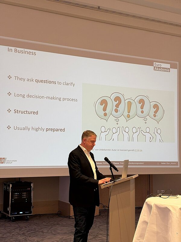 Ein Mann im Anzug steht an einem Rednerpult und hält einen Vortrag. Im Hintergrund ist eine Präsentationsfolie projiziert, die den Titel 'In Business' trägt. Die Folie enthält vier Aufzählungspunkte und eine farbige Illustration mit fünf Personen, die jeweils ein Schild mit einem großen Fragezeichen hochhalten. In der rechten oberen Ecke der Folie befindet sich ein rot-weißes Logo mit der Aufschrift 'Euro Akademia'. Der Raum wirkt modern und ist mit Technik ausgestattet.