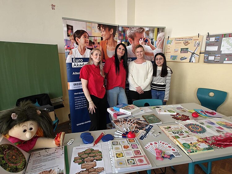 Vier lächelnde Personen posieren vor einem Roll-up-Banner mit der Aufschrift 'Euro Akademie' und dem Hinweis auf Erziehung und Bildung. Auf dem Tisch vor ihnen sind verschiedene pädagogische Materialien wie Bücher, Bastelvorlagen, Stifte und Informationsblätter ausgelegt. Im Vordergrund sitzt eine große Stoffpuppe mit braunen zotteligen Haaren.