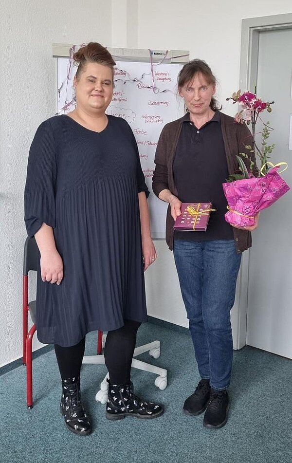 „Pflege und Betreuung ist Arbeit mit dem Herzen“ Zwei Frauen stehen nebeneinander in einem Büro. Die Frau links trägt ein dunkelblaues Kleid und schwarze Stiefel mit weißen Mustern. Die Frau rechts hält einen Blumentopf mit einer Orchidee und ein verpacktes Geschenk in den Händen. Sie trägt eine dunkle Hose, ein schwarzes Polohemd und eine braune Strickjacke. Im Hintergrund befindet sich ein Flipchart mit handschriftlichen Notizen auf Deutsch.