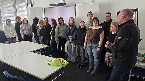 Eine Gruppe von jungen Auszubildenden der Euro Akademie Erfurt steht in einem Raum mit weißen Wänden und mehreren Tischen. Sie alle sind in lockerer Formation aufgestellt und blicken in Richtung eines Mannes, der ihnen gegenübersteht. Er trägt eine dunkle Jacke und hält die Hände vor dem Körper. Im Hintergrund sind ein Whiteboard, eine Uhr und Fenster mit Jalousien zu sehen. Auf einem Tisch liegt eine gelbe Tasche.